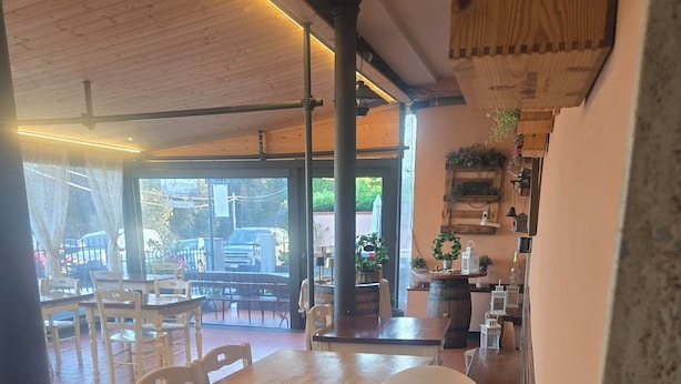 Visão interna de um restaurante com teto de madeira, grandes janelas e área de refeições com mesas e cadeiras.