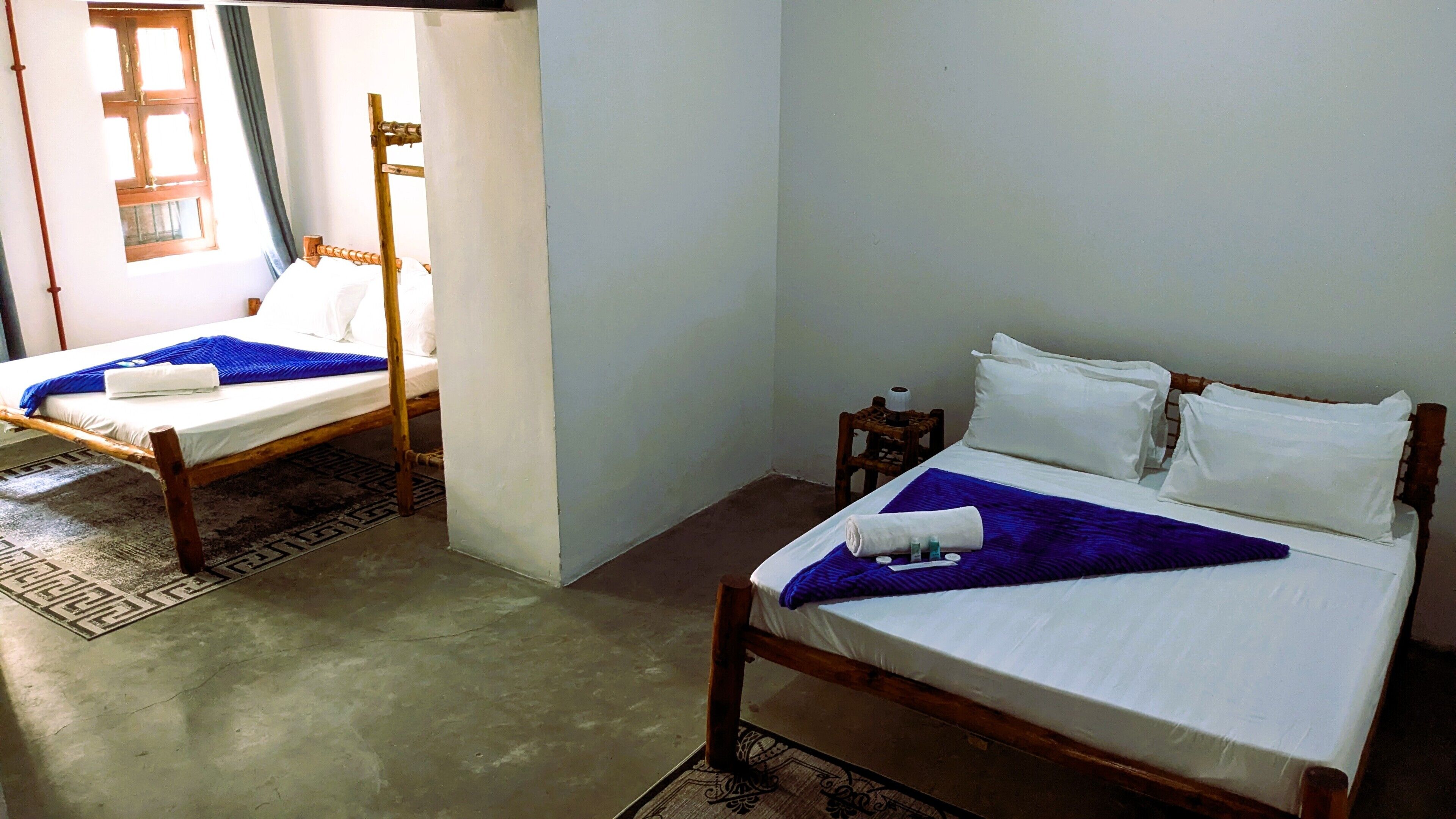 Deux lits en bois avec du linge de lit blanc, une petite table en bois avec des articles de toilette et un tapis à motifs.