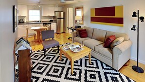 Ein Wohnzimmer mit einer Couch, einem Couchtisch, einem gemusterten Teppich, einem Essbereich mit Stühlen und einer Küche im Hintergrund.