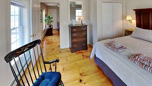 Ein Schlafzimmer mit einem Bett, einem Schminktisch, einem Sessel, einem Deckenventilator und einem Flur, der in einen weiteren Raum führt.