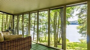 En inglasad veranda med en flätstol och ett litet bord, med utsikt över en sjö som omges av träd.
