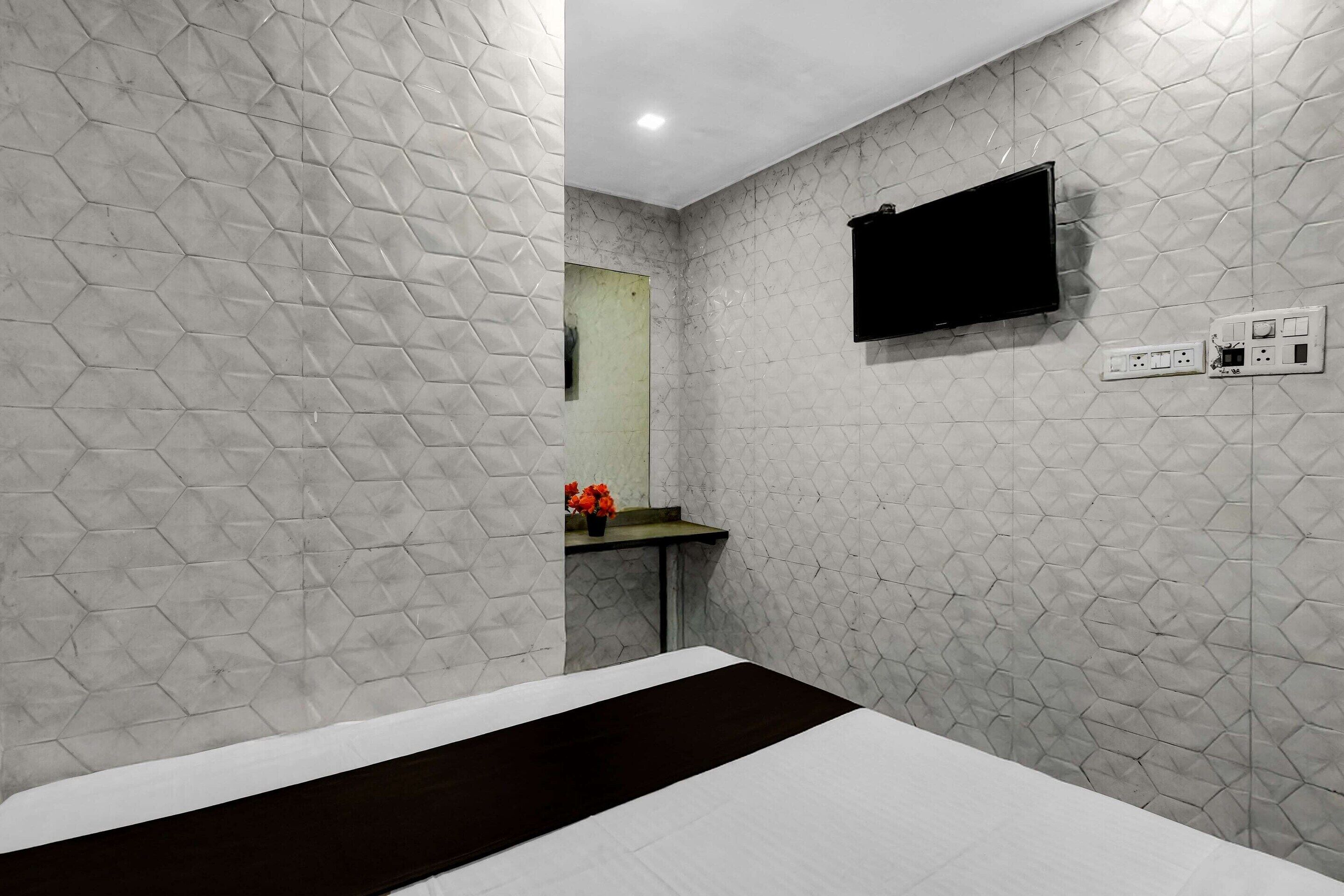 A hotel room with a bed, a flat-screen TV mounted on the wall, a small table with a vase of flowers, and a mirror.