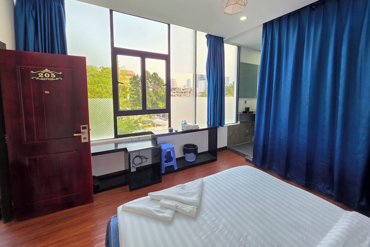 Une chambre d’hôtel avec un grand lit, un bureau avec une chaise, une fenêtre avec des rideaux, une porte avec une plaque numérotée et une vue sur l’extérieur.