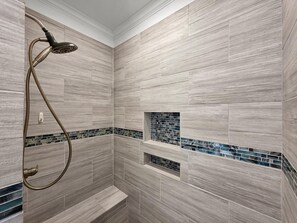 A shower with a handheld showerhead and a built-in seat, tiled walls with a wood-like pattern, and a small decorative tile accent. - Heaven's Song 3 Bedroom Home (Port Bolivar)