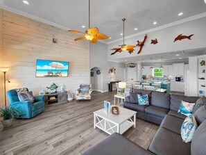 A spacious living area with a wooden floor, a ceiling fan, a television, and a dining area with a kitchen in the background. - Heaven's Song 3 Bedroom Home (Port Bolivar)