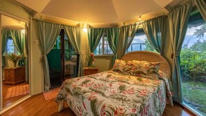 Ein Schlafzimmer mit Bett, Vorhängen, einem Nachttisch, einer Vase mit Blumen und Blick auf Grün.