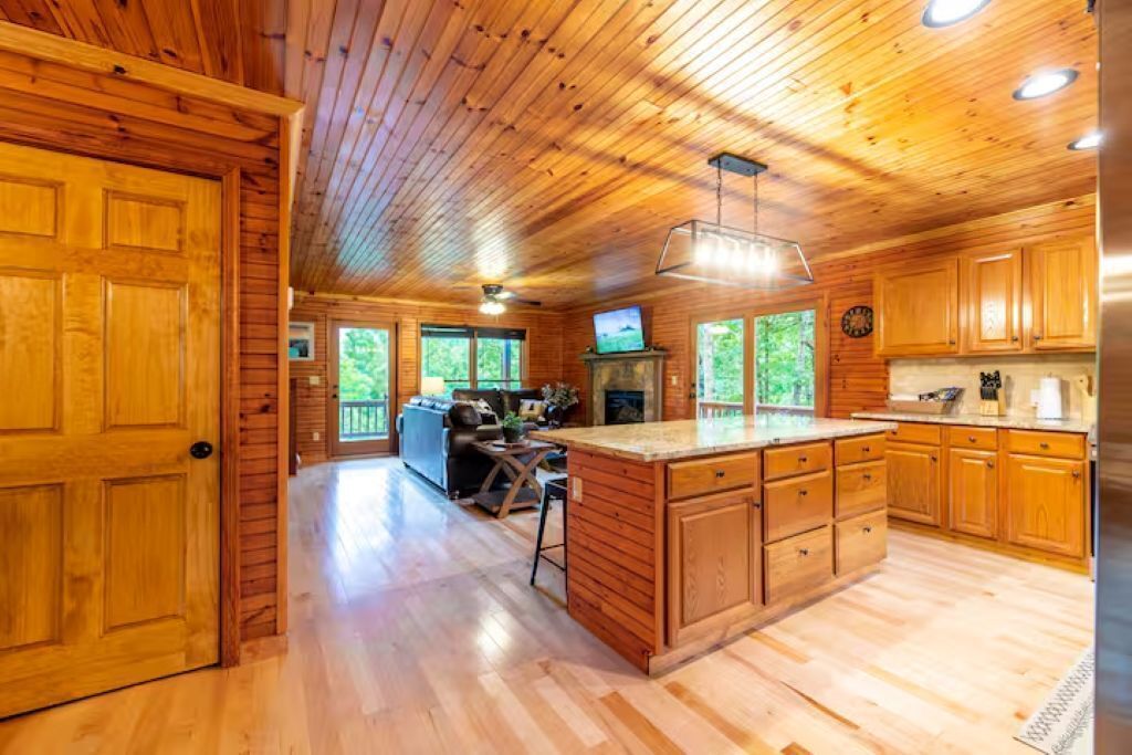 A cabin interior with wooden walls, a kitchen with wooden cabinets, a dining area, and a fireplace.