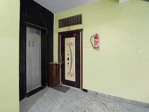 A hallway with a fire extinguisher mounted on the wall, a door with a number plate, and a small trash bin. - Deluxerajgir Junction Hotel Mamta Kutir (Rajgir)
