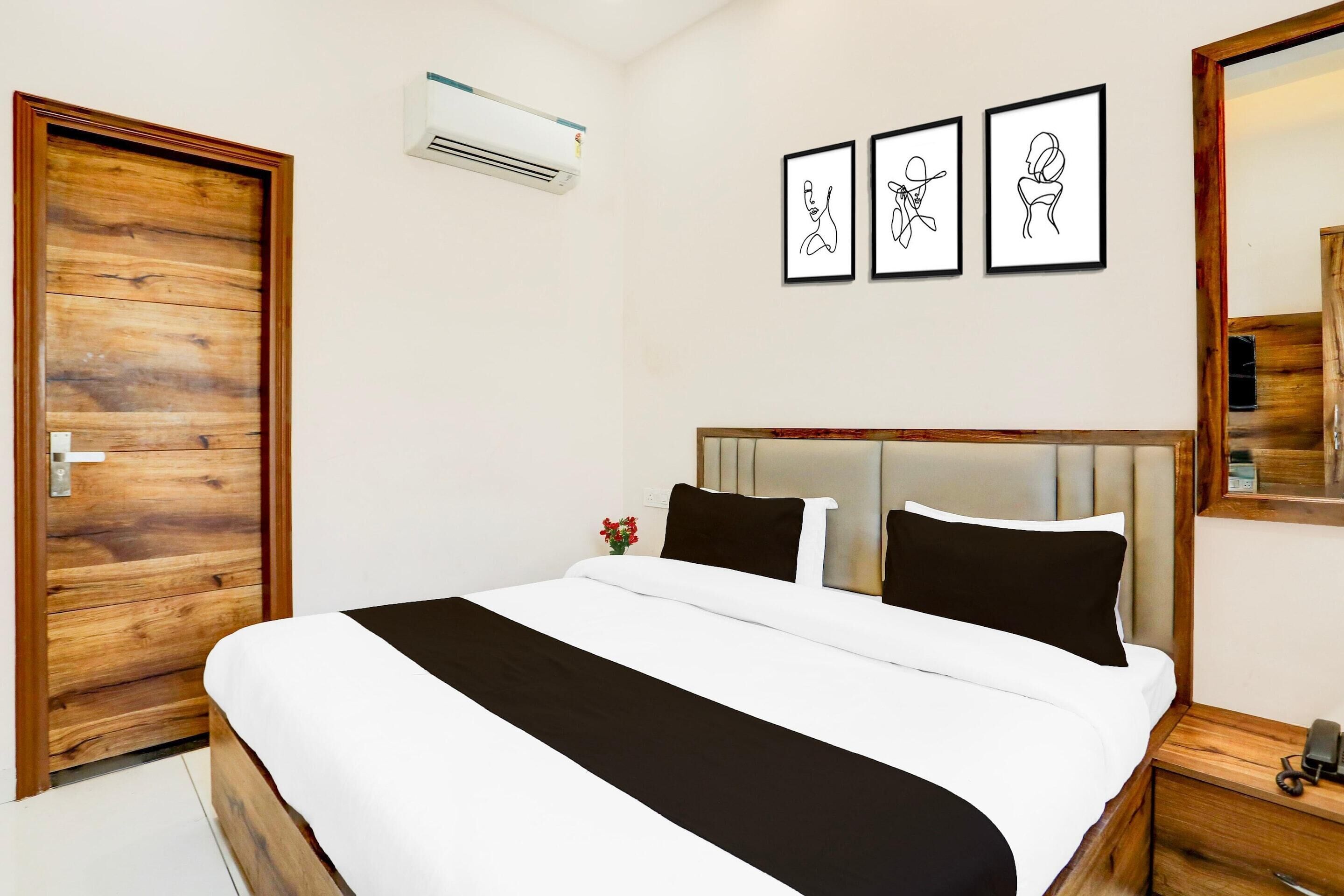 Een hotelkamer met een houten deur, een bed met een zwart-wit dekbedovertrek, een nachtkastje met een telefoon en drie ingelijste kunstwerken aan de muur.