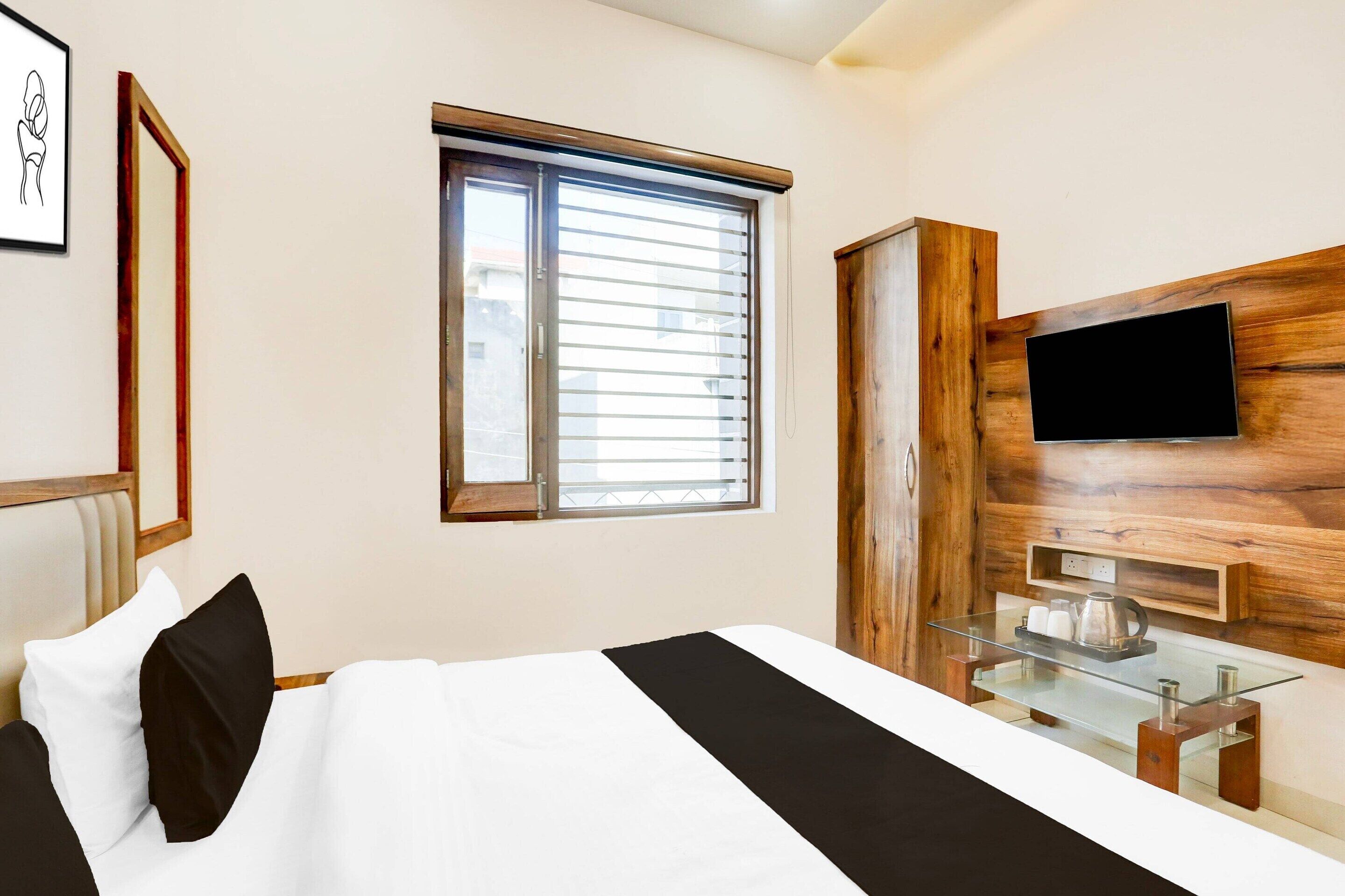 A modern hotel room with a bed, a TV mounted on a wooden wall, a window with blinds, and a glass coffee table.