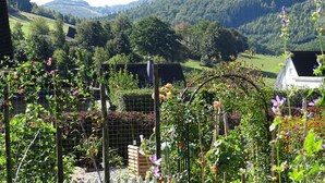 A garden with various plants and flowers, a wooden fence, and a view of a hill with a house in the background. - Anno 1908 - Holiday home in the Sauerland (Olsberg)