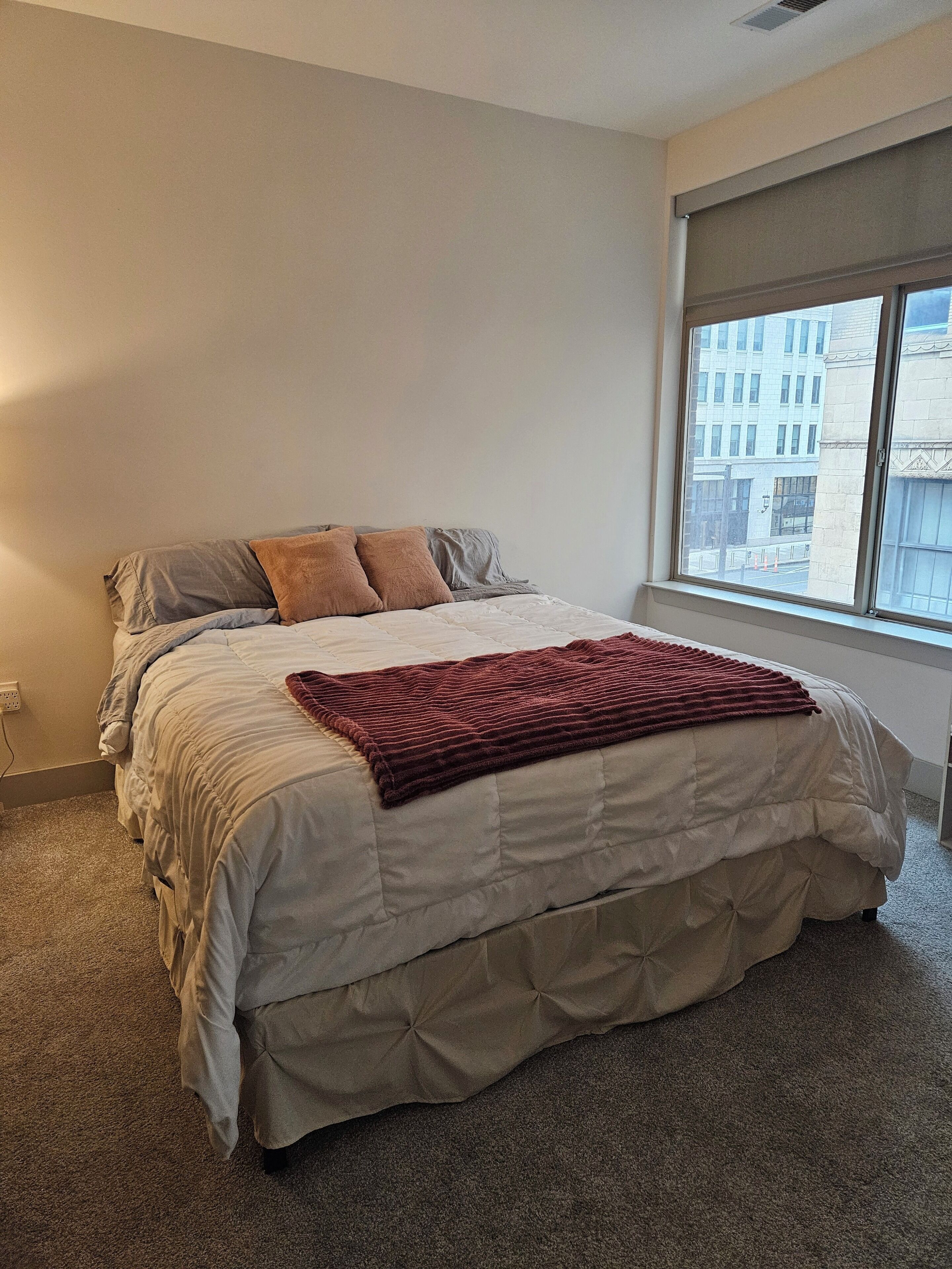 Une chambre avec un grand lit, une fenêtre donnant sur des immeubles et un sol recouvert de moquette.