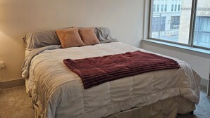 A bedroom with a large bed, a window with a view of buildings, and a carpeted floor.