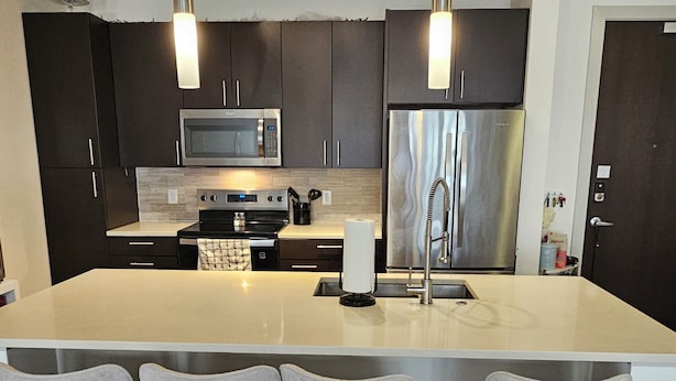 A modern kitchen with dark cabinets, stainless steel appliances, a large island with bar stools, and pendant lighting.