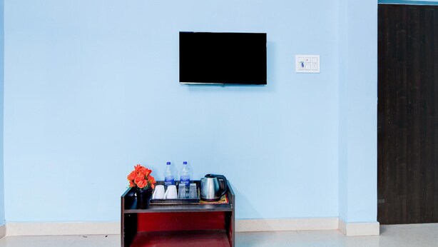 Ein Zimmer mit einem an der Wand befestigten Flachbildfernseher, einem kleinen Tisch mit Tablett, auf dem Wasserflaschen, Becher und ein Wasserkocher stehen, sowie einer Vase mit Blumen.