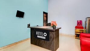 Ein Rezeptionstresen mit der Aufschrift 'HOTEL', einem Schild mit 'Company-Served', einem Flachbildfernseher, einer Zimmerpflanze und einem roten Sessel.