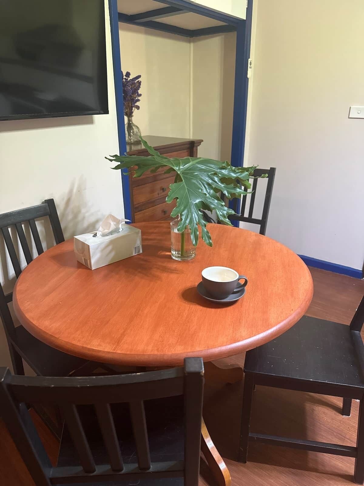 A round wooden dining table with chairs, a vase with a plant, a tissue box, and a cup of coffee.