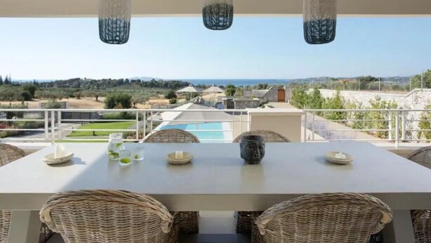 Eine Terrasse im Freien mit Tisch, Korbstühlen, hängenden Pendelleuchten und Blick auf einen Pool und die Landschaft.