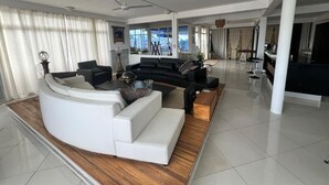 A modern living room with a curved white sofa, a black sectional sofa, and a wooden coffee table. There is a ceiling fan, a kitchen area in the background, and large windows with curtains. - Penthouse 3 min walk from Flic en Flac beach (Flic en Flac)