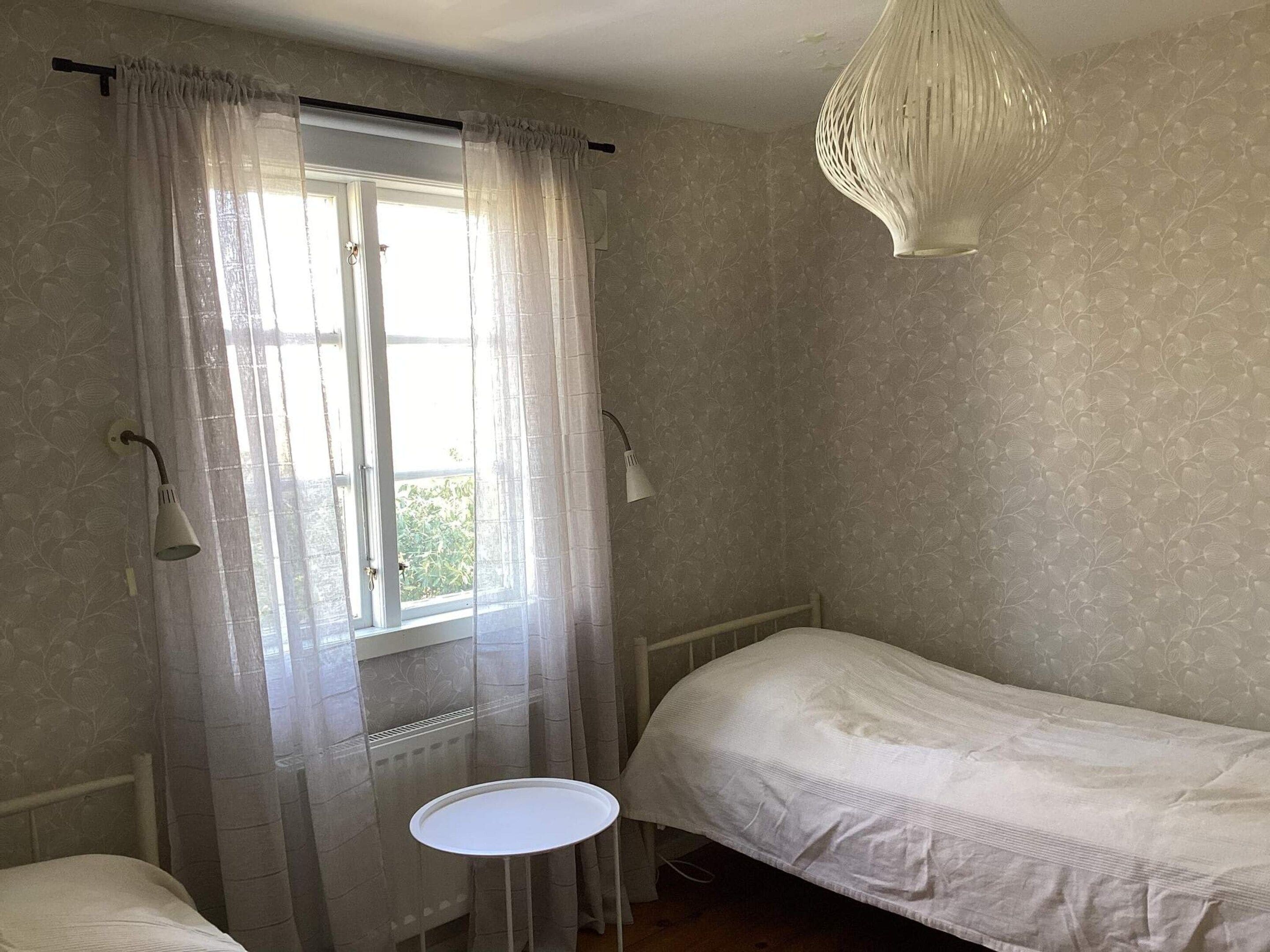 Un dormitorio con una cama individual, una mesita redonda pequeña, una ventana con cortinas traslúcidas y una lámpara de pared.