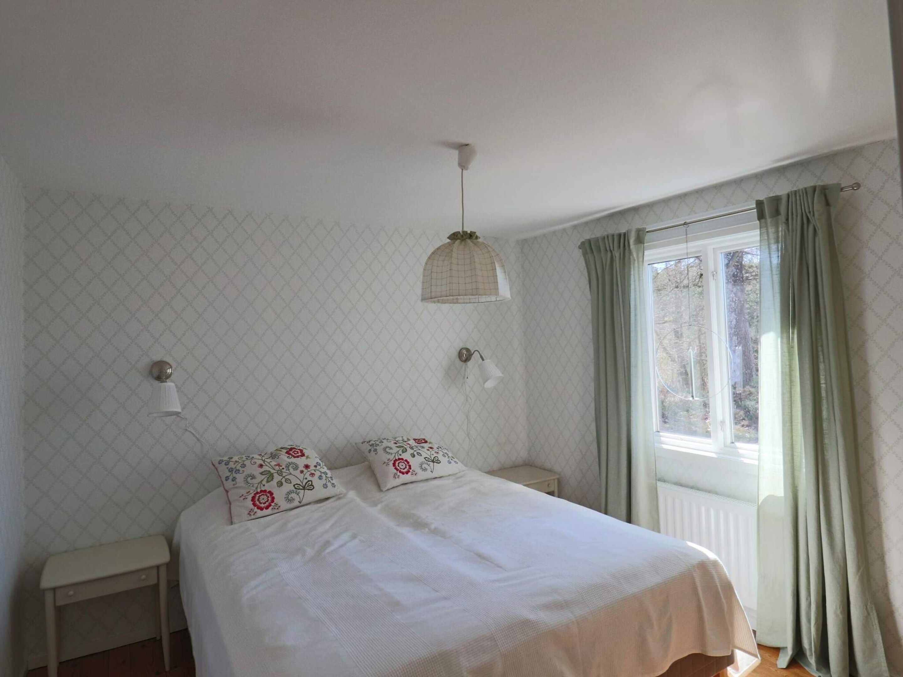 Un dormitorio con cama, dos mesitas de noche, una lámpara colgante y una ventana con cortinas.
