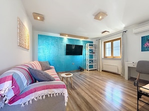 A living room with a blue accent wall, a sofa with striped cushions, a small round table, a white bookshelf, a TV mounted on the wall, and a window with curtains. - Apartment Adri by Interhome (Rovinj)