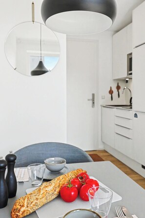 Une cuisine moderne avec des meubles blancs, un miroir rond et une table à manger sur laquelle on trouve une miche de pain, des tomates et des couverts.