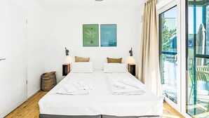 Cama doble con sábanas blancas, dos lámparas fijadas a la pared, una mesita pequeña y dos cuadros enmarcados de Berlín y Copenhague.