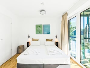 Ein Doppelbett mit weißen Leinentüchern, zwei Wandlampen, ein kleiner Nachttisch und zwei gerahmte Bilder von Berlin und Kopenhagen.