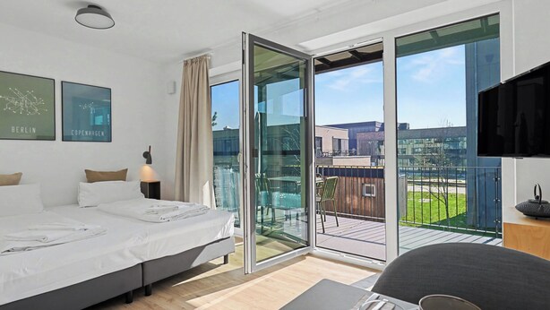 Una habitación de hotel moderna con una cama, una mesa de comedor con un tazón y cubiertos, un televisor y un balcón con vistas a la ciudad.