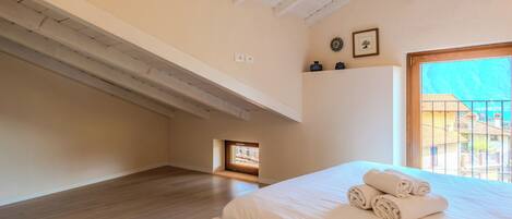 Quarto com piso de madeira, cama com roupa de cama branca, claraboia e vista para edifícios.