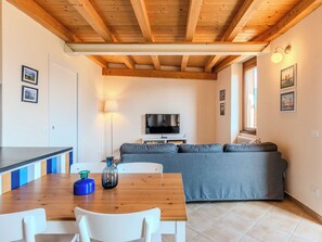 Una sala de estar con techo de madera, un sofá, un televisor, una mesa de comedor con sillas y un ventanal.