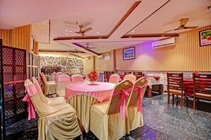 Un salón de banquetes con mesas redondas, sillas con cintas rosas, ventiladores de techo y un equipo de aire acondicionado.