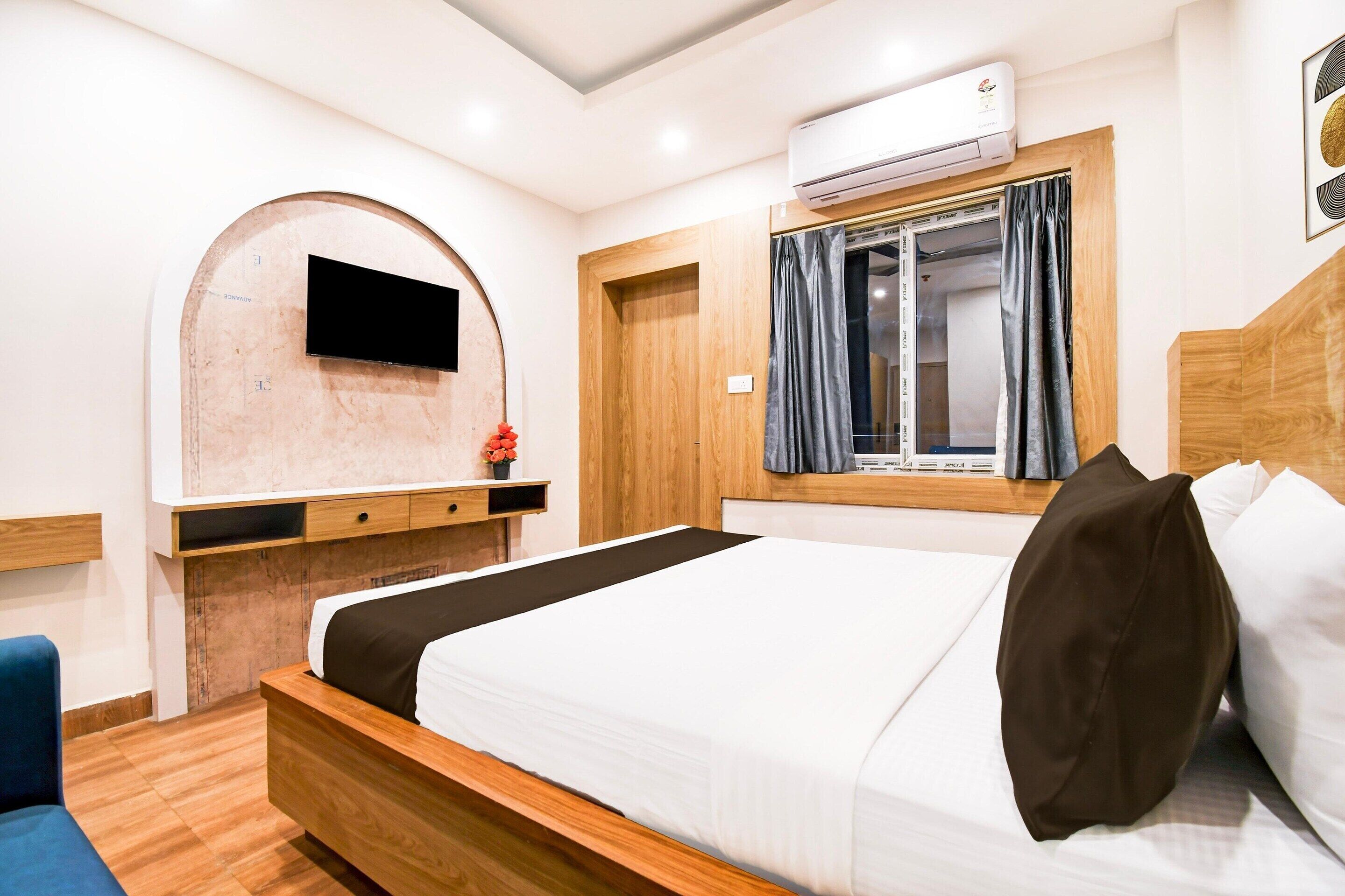 Ein modernes Hotelzimmer mit einem hölzernen Bett, einem an der Wand befestigten Flachbildfernseher, einem Fenster mit Vorhängen und einem eingebauten Kleiderschrank.