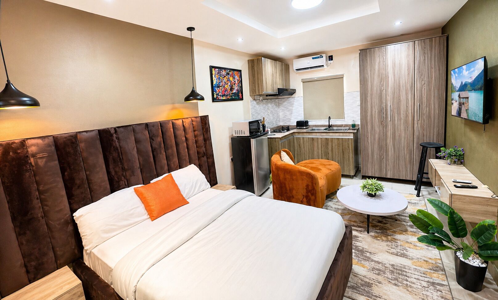 Habitación de hotel con cama, un pequeño sofá, una mesa redonda, una planta en maceta, televisor y una kitchenette.