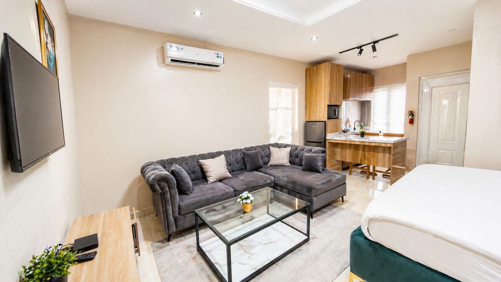 A modern living room with a sectional sofa, a glass coffee table, a TV mounted on the wall, a kitchenette with wooden cabinets, and a bed with white bedding.
