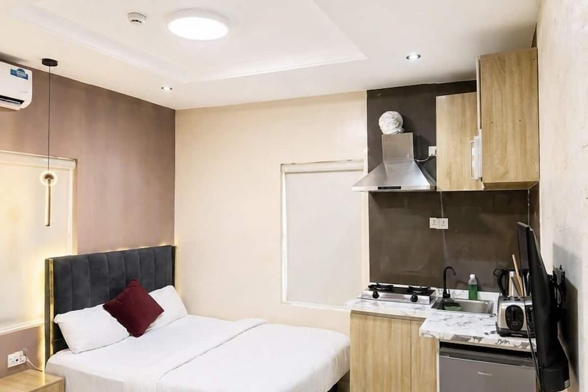 Quarto de hotel moderno com cama, mesa de cabeceira, escrivaninha, cozinha compacta e um pequeno espaço de estar.