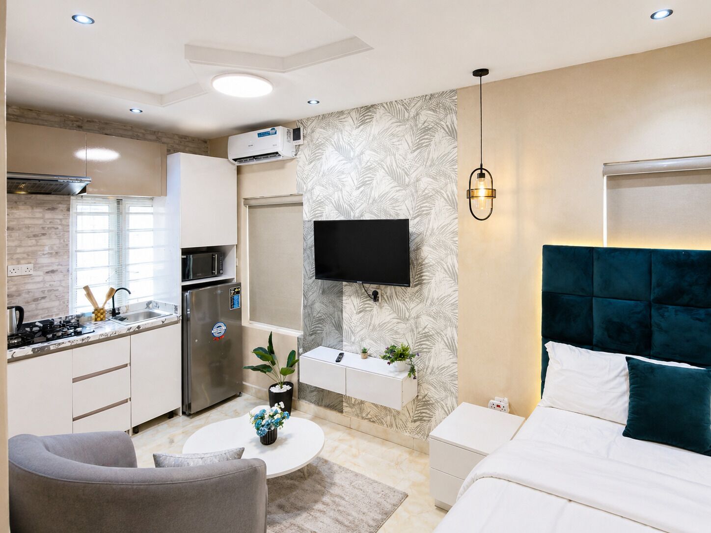 A compact hotel room with a kitchenette, a flat-screen TV mounted on the wall, a bed with a teal headboard, and a small seating area.