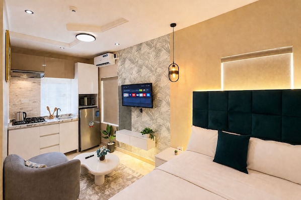 A modern hotel room with a bed, a TV, a kitchen area, and a small seating area.