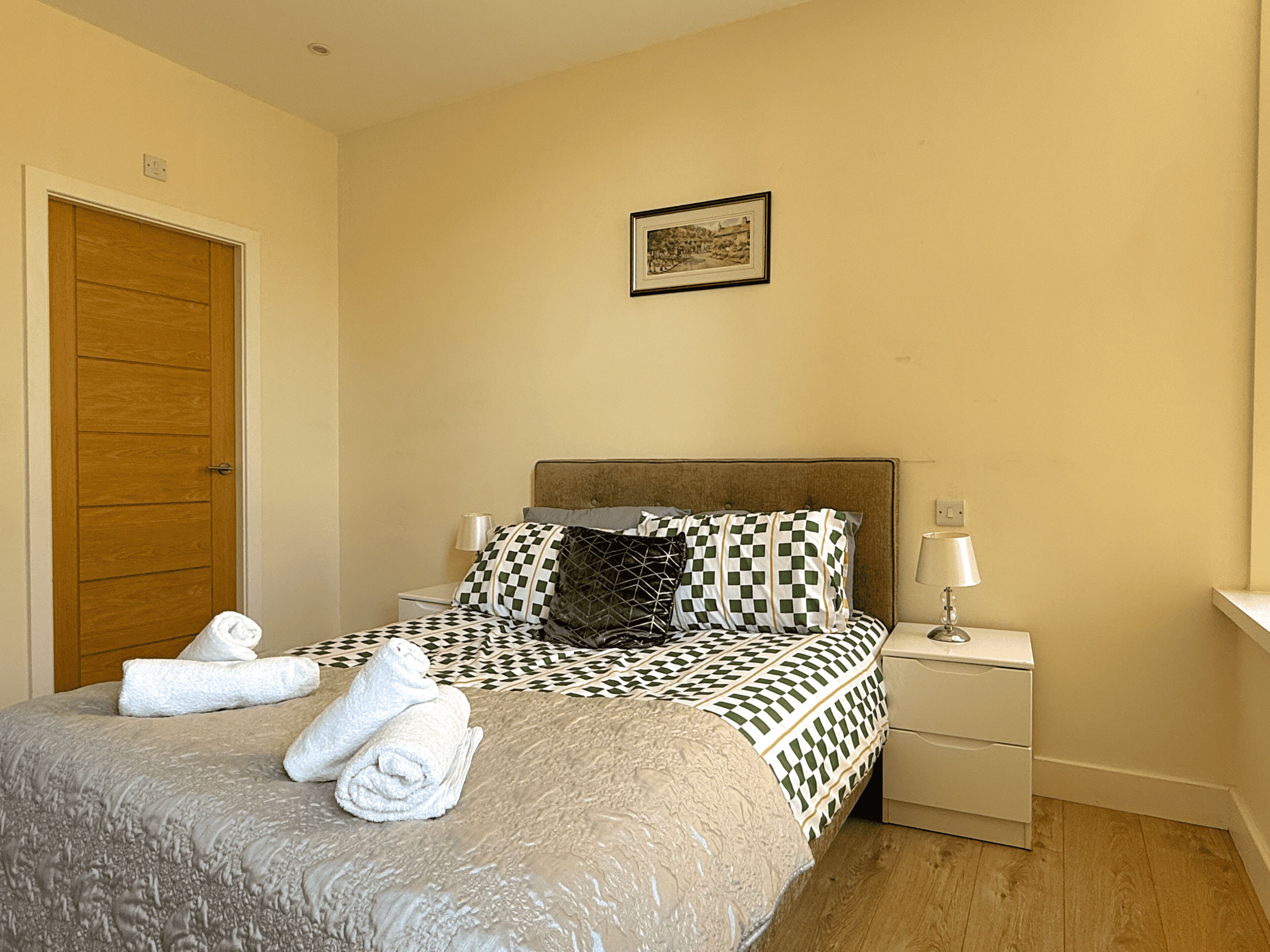 A neatly made bed with a patterned bedspread, two pillows, and rolled towels on the side. There is a nightstand with a lamp, a framed picture on the wall, and a wooden door.