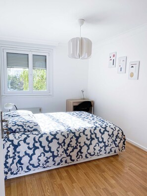 Een slaapkamer met een bed, een raam met zonwering, een nachtkastje, een lamp en een kunstwerk aan de muur.