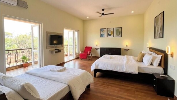 A hotel room with two beds, a TV, a balcony with a potted plant, and artwork on the wall. - 4-bedroom house in delightful Coimbatore with cool AC and space (Coimbatore)