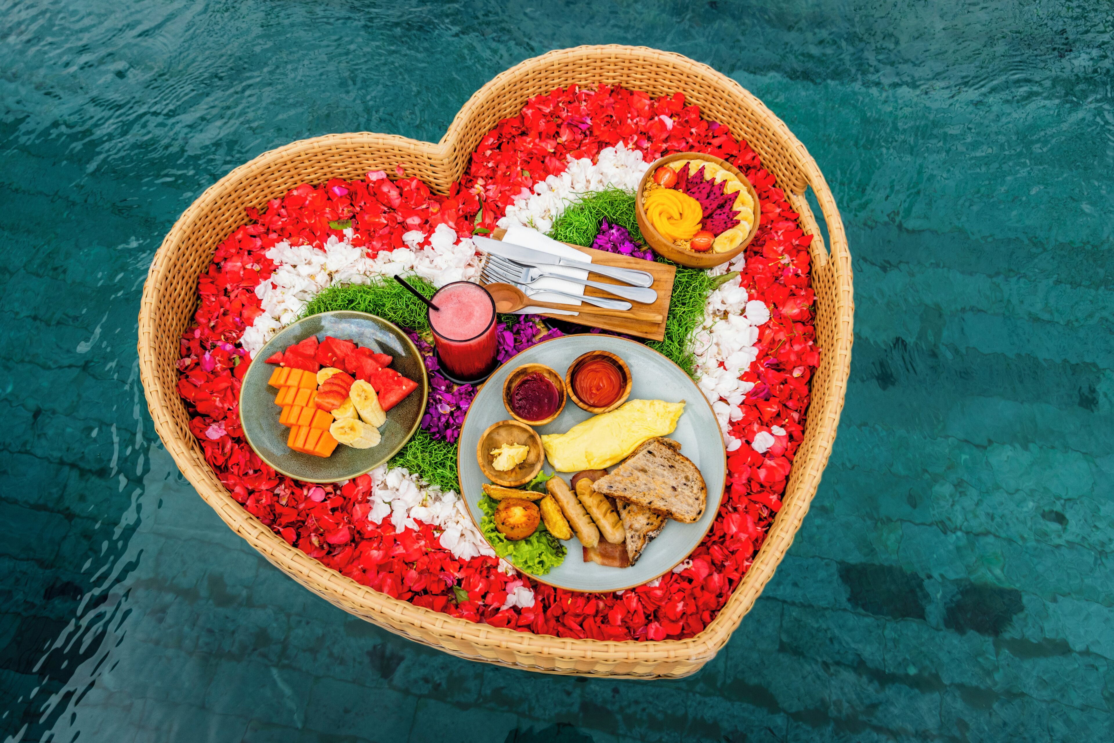 A heart-shaped basket filled with food items and utensils, placed on a body of water.