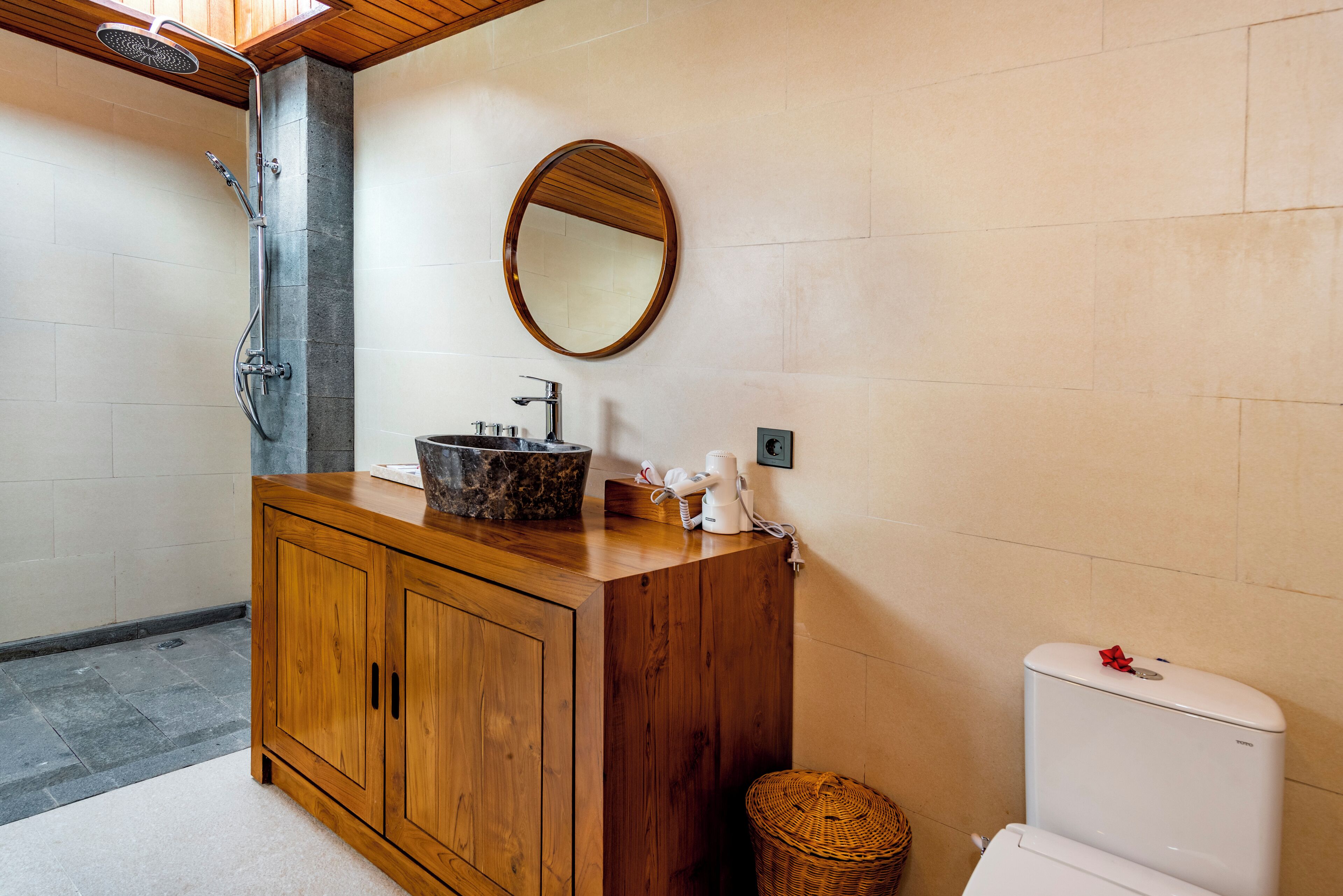 Un baño con tocador de madera, espejo redondo, lavamanos, ducha e inodoro.