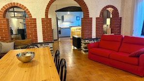 A living room with a red sofa, a wooden dining table with a metal bowl, and brick arches. - A peaceful stay among the olive trees (Virgili)