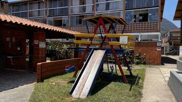 Um parquinho com escorrega e balanços, um edifício de tijolos com telhado de telhas, e um prédio residencial de vários andares ao fundo.