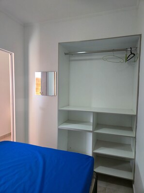 A room with a bed covered in blue, a white wardrobe with multiple shelves, and a mirror on the wall.