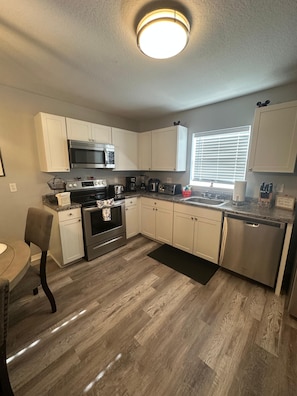 A kitchen with white cabinets, stainless steel appliances, a window with blinds, and a dining area with a chair. - Cozy family home minutes from Disney, fully equipped with Wi-Fi, pool… (Kissimmee)