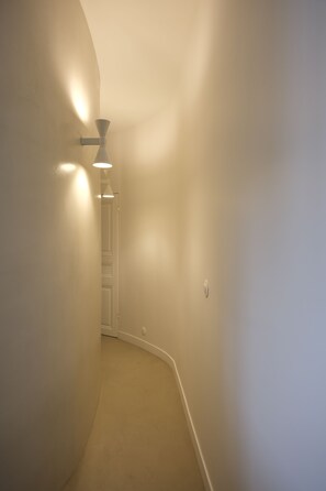 Ein schmaler Flur mit einer Tür am Ende, einer Wandleuchte und einem Lichtschalter an der Wand.