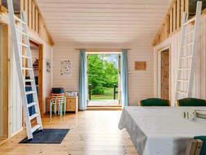 En stuga med träpanel på insidan, med ett matbord, stolar, en stege och utsikt över grönskan utanför.